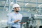 Man stands in the production area wearing a white hard hat and holding a laptop.