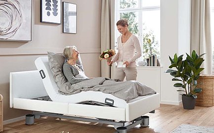 Ältere Frau liegt im Bett und bekommt von einem Besuch Blumen.