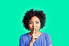 Woman with blue shirt holds finger in front of mouth and whispers.