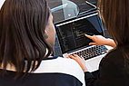 Zwei Frauen sitzen vor einem Laptop und erklären sich etwas.
