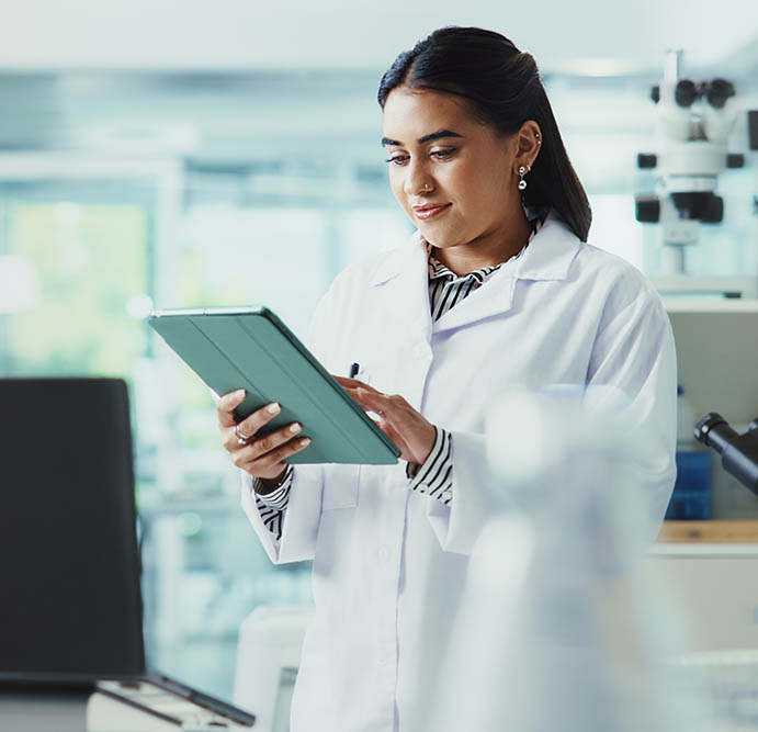 Wissenschaftlerin im Labor trägt einen Kittel und nutzt ein Tablet zur Datenerfassung oder Analyse neben einem Mikroskop.